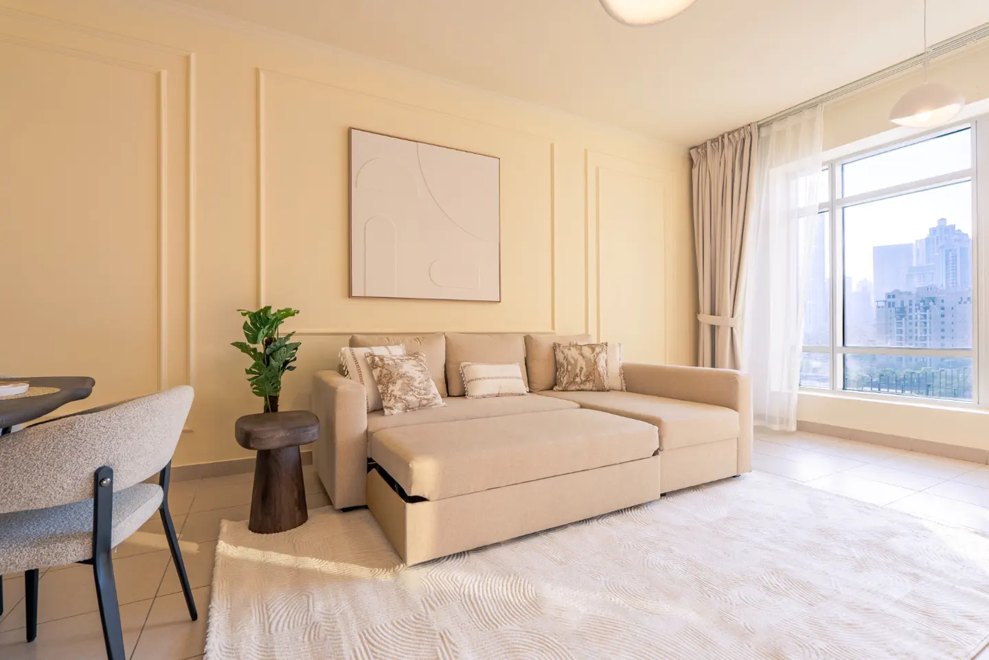Living room with a beige pull-out sofa, decorative pillows, and a white rug. A plant sits on a wooden stool. Large window with city view.