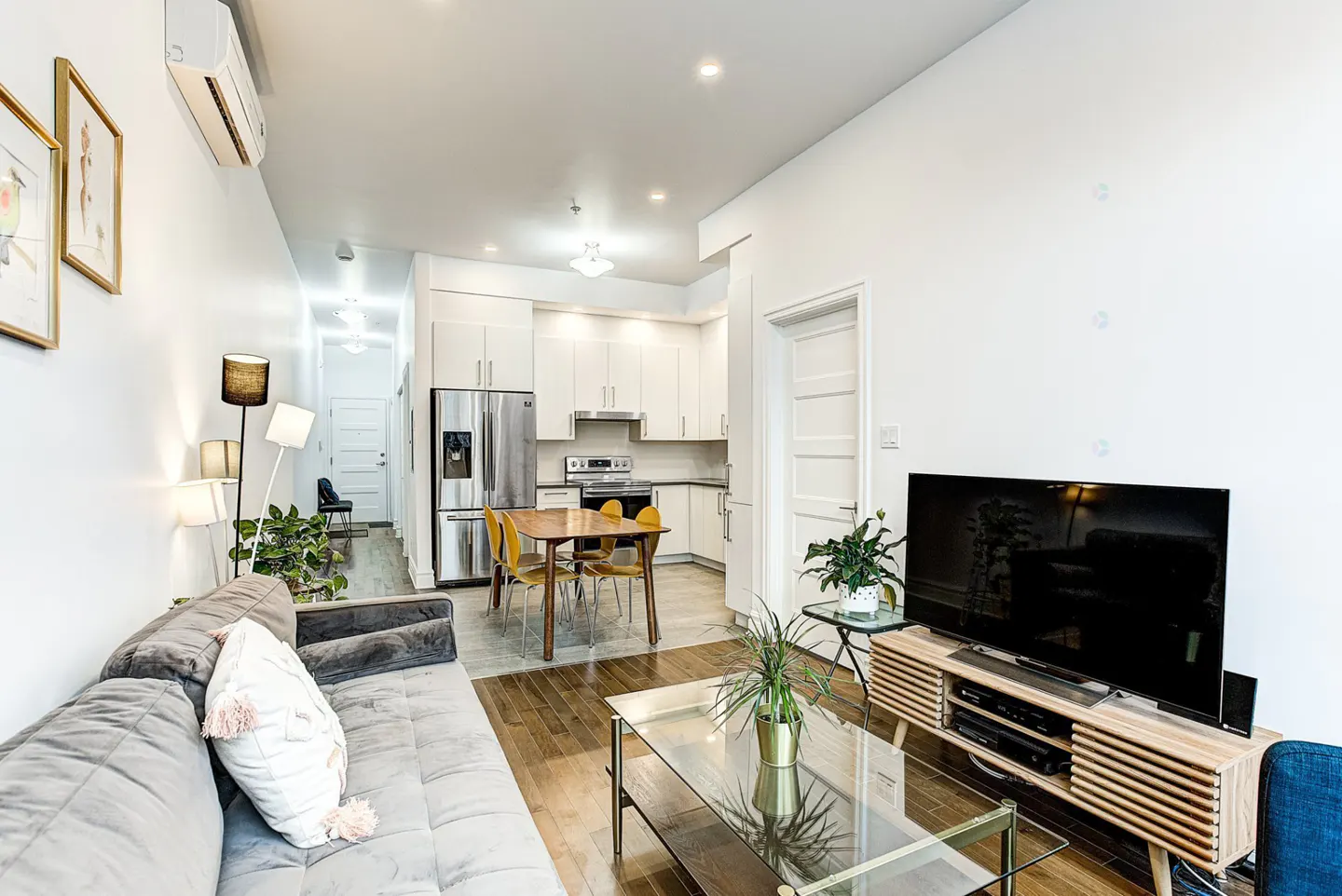 Modern open-concept living room with a grey sofa, glass coffee table, TV console, and a kitchen with white cabinets.