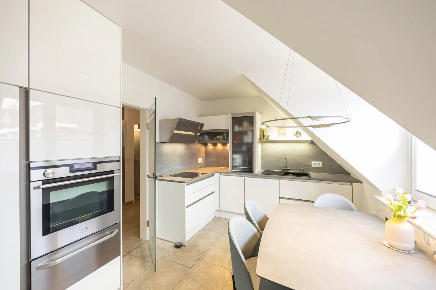 Bright, modern kitchen with white cabinets, stainless steel oven, and dining table with grey chairs. A vase of tulips sits by the window.
