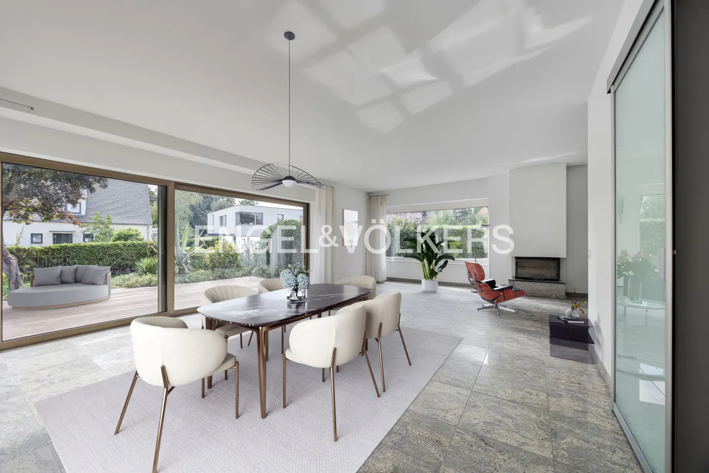 Bright, modern dining room with a marble table, cream chairs, and a large window overlooking a green yard. A fireplace and red chair are visible in the background.