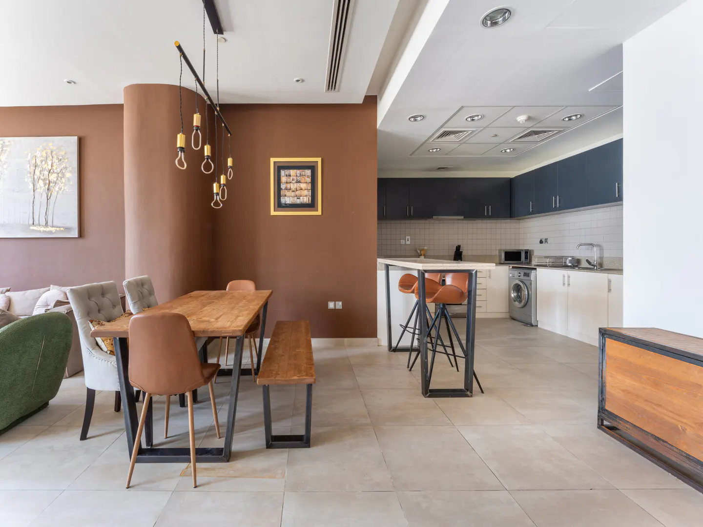 A modern apartment interior with a dining area featuring a wooden table, chairs, and a bench, adjacent to a kitchen with blue and white cabinets.