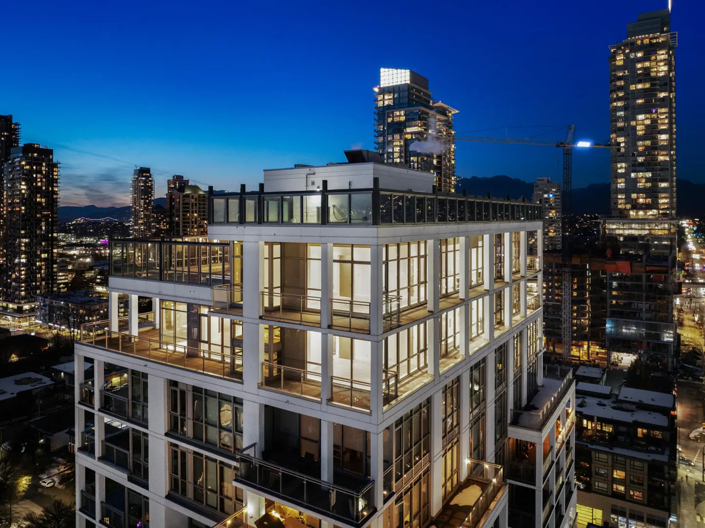 Aerial view of a modern white high-rise apartment building at twilight, surrounded by city lights and other skyscrapers.