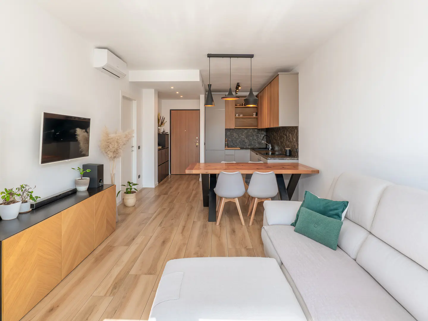 Bright, modern apartment interior with white sofa, wood floors, and open kitchen. Black pendant lights hang above a wooden table with gray chairs.