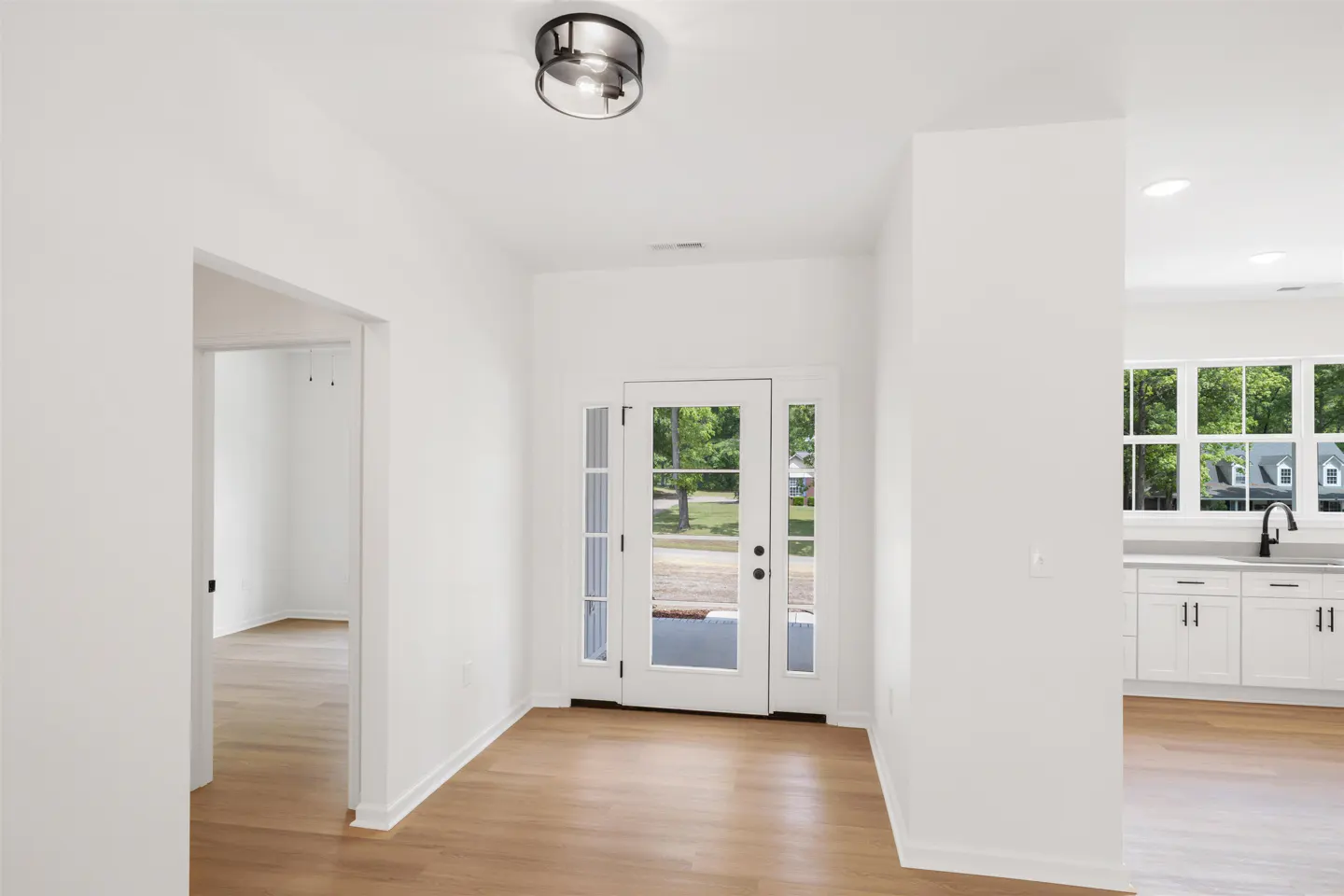 Bright, white entryway with wood floors and a glass-paneled front door. A glimpse of a kitchen with white cabinets is visible.