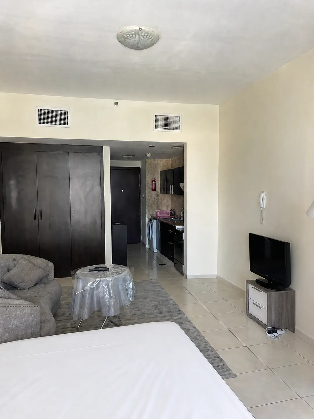 A bright studio apartment with a bed, sofa, TV, kitchen, and dark wood cabinets.