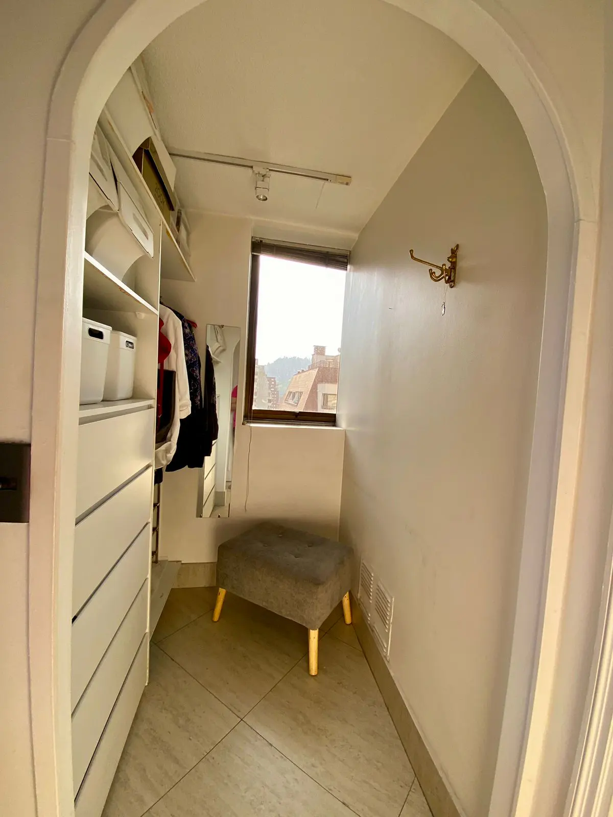 Walk-in closet with white shelving, drawers, and a gray ottoman. A window offers a view of buildings and hills.