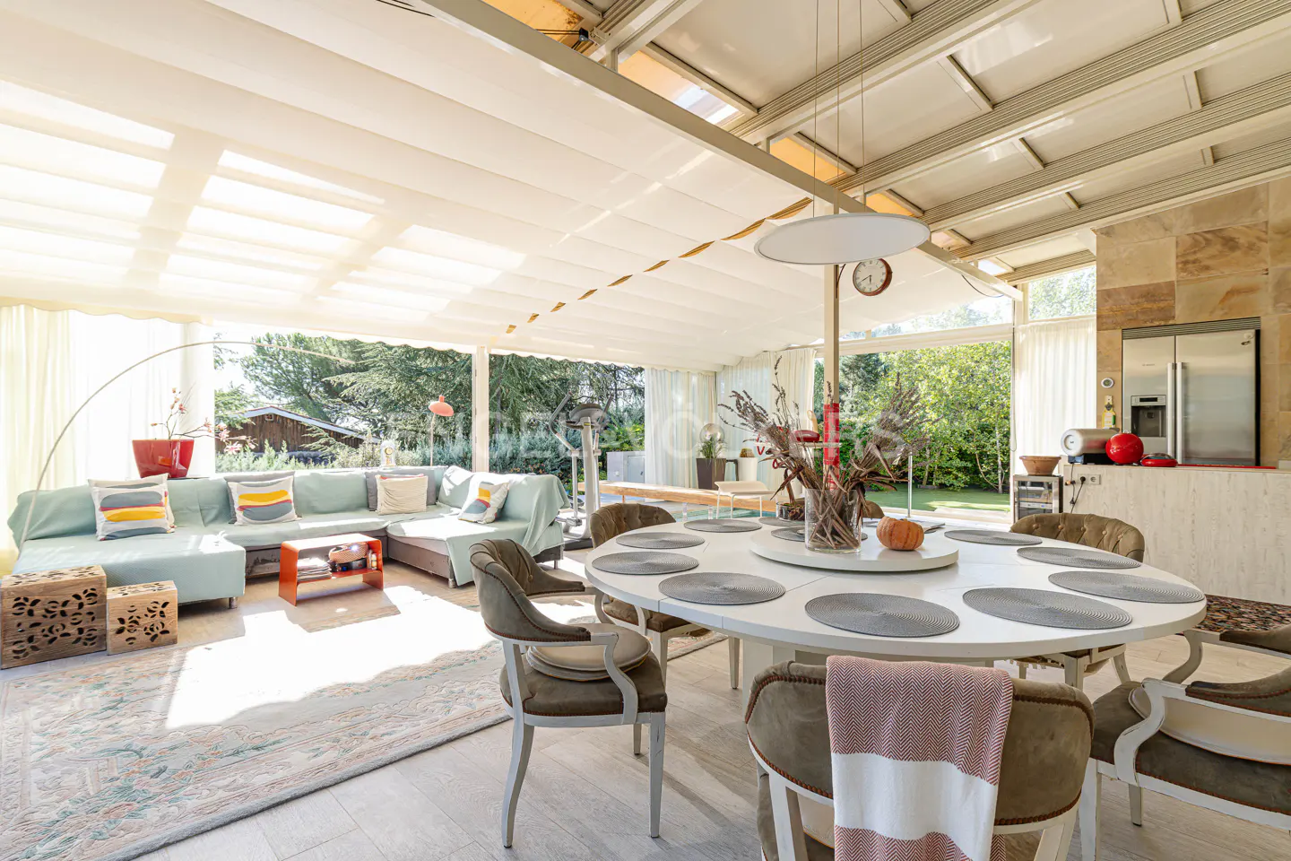 Bright sunroom with a white dining table, olive chairs, and a light blue sectional sofa. Large windows overlook a green yard.