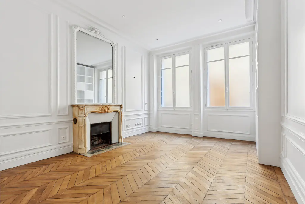 Bright, empty room with herringbone wood floors, white walls, fireplace with mirror, and two large windows.