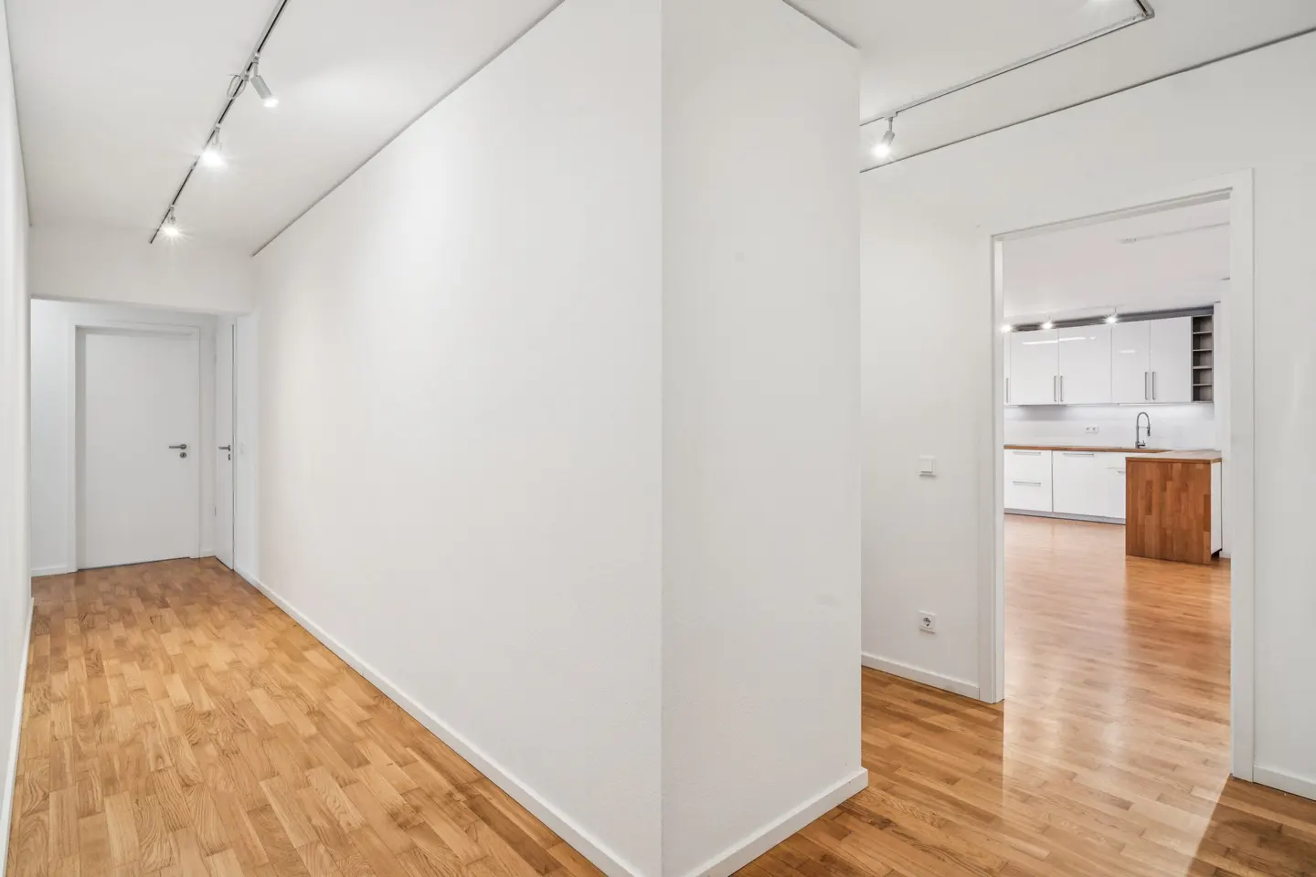 Hallway with hardwood floors and white walls. A kitchen is visible through a doorway. Track lighting on the ceiling.
