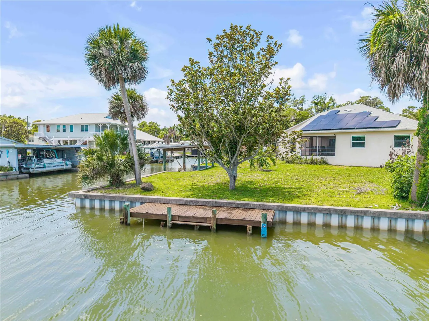 Waterfront home with a wooden dock on a canal. Palm trees and green grass surround the white house with solar panels.