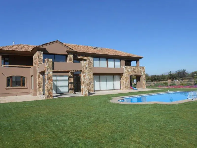 Two-story brown house with stone accents, a tile roof, and a blue swimming pool on a green lawn under a clear blue sky.