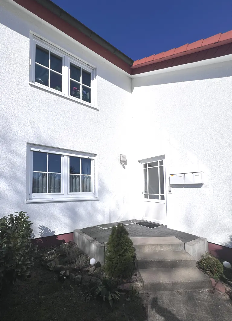Exterior of a white house with a red roof, two windows, a door, and a small garden.