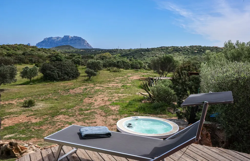 Outdoor spa with a lounge chair and hot tub on a wooden deck, overlooking a green landscape and distant mountain under a blue sky.
