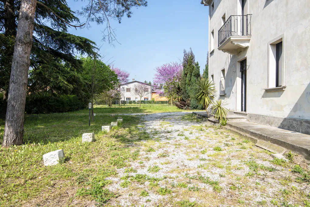 Exterior view of a light-colored two-story house with a balcony, surrounded by green trees and grass on a sunny day.