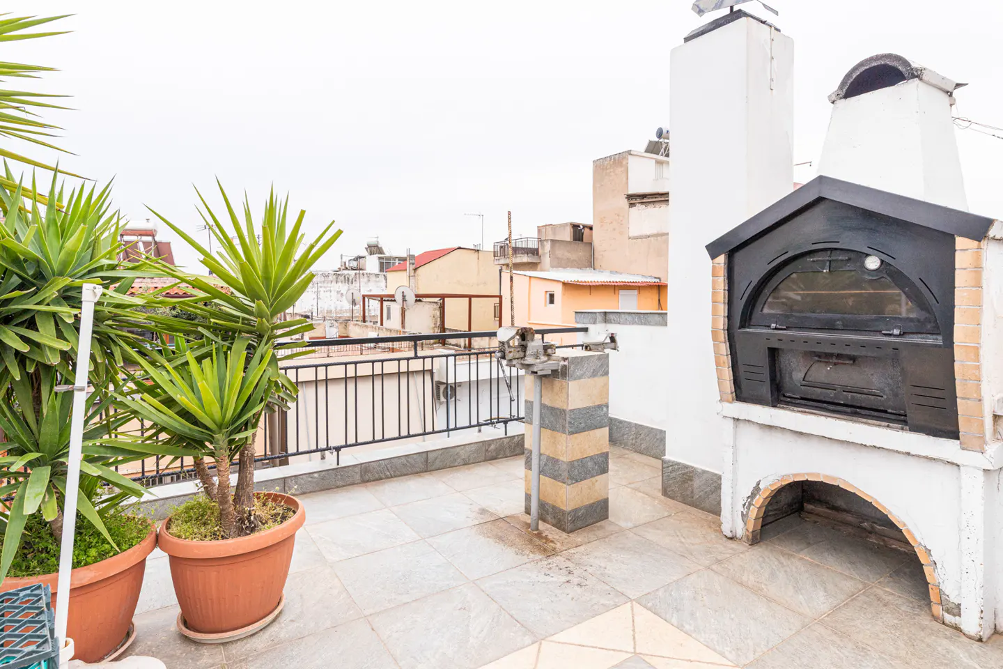 Rooftop terrace with potted plants, grill, and brick oven. City buildings are visible in the background.