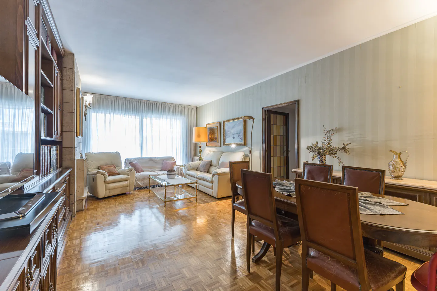 Bright living and dining room with parquet floors. Cream sofas, brown leather chairs, and a large wooden bookcase.