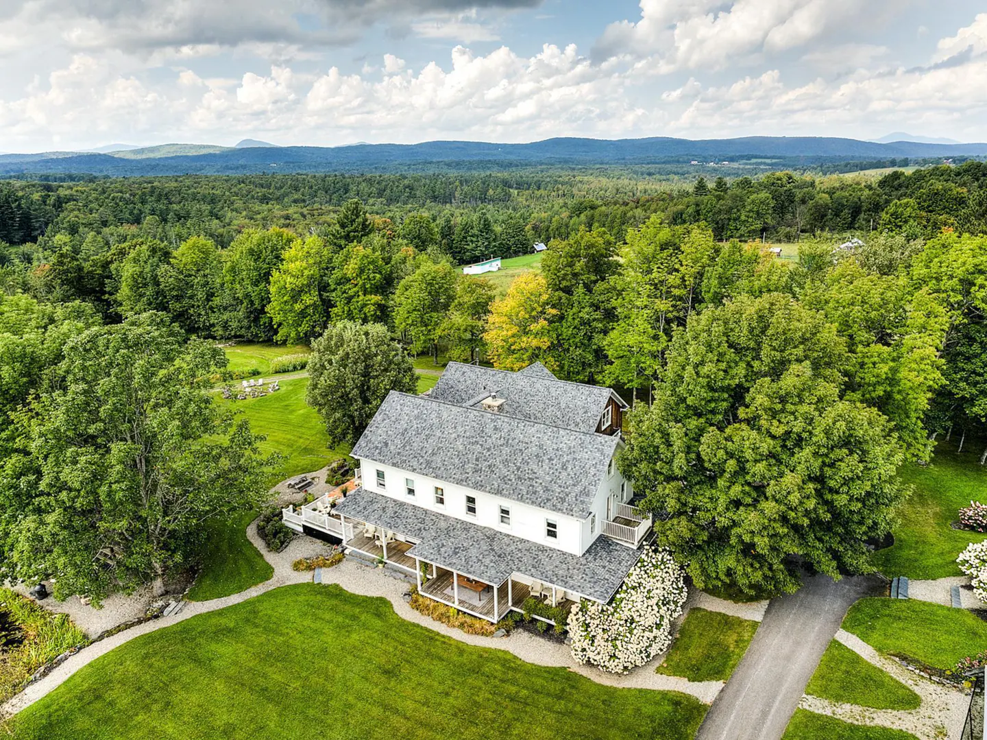 Aerial view of a white farmhouse with a gray roof, surrounded by green lawns, trees, and distant mountains under a cloudy sky.