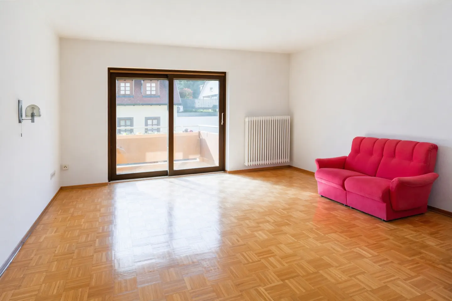 Bright, empty room with parquet floor, white walls, radiator, and a red sofa. Sliding glass doors lead to a balcony.