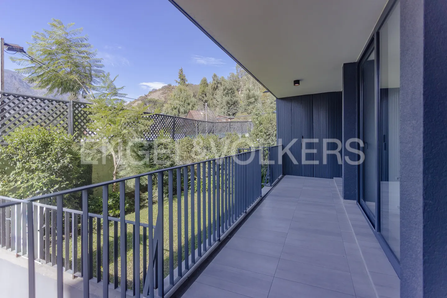 A balcony with gray railings overlooks a green lawn and trees under a blue sky. The balcony has gray tile flooring and a dark gray exterior wall.