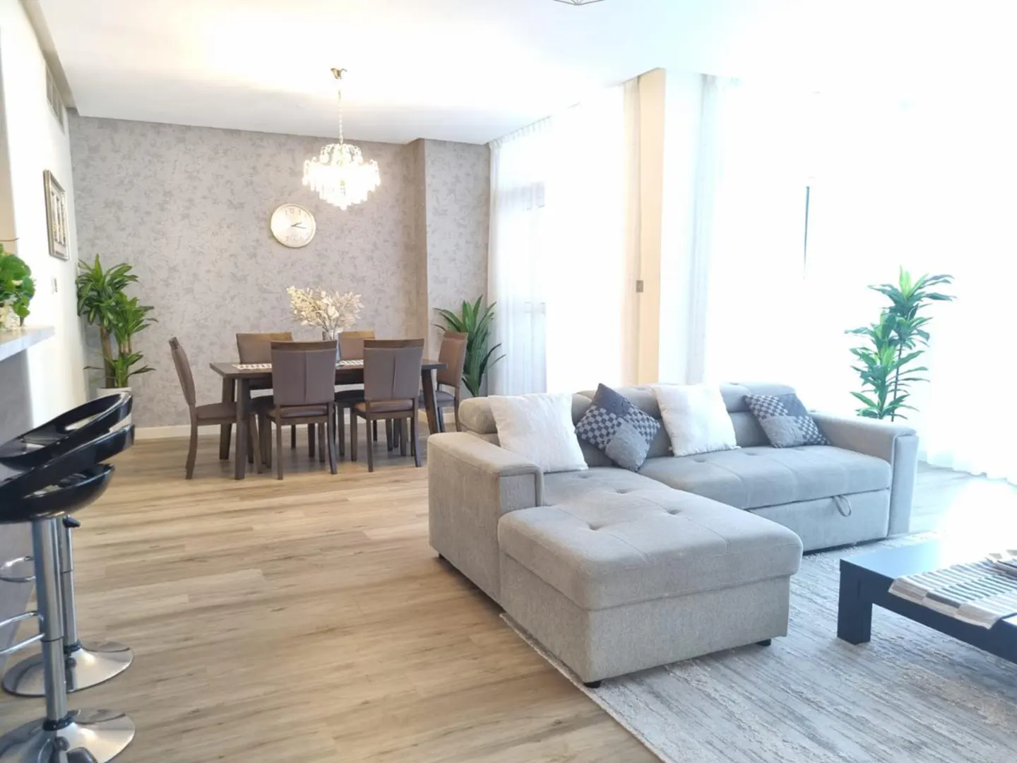 Bright, open-plan living space with a gray sectional sofa, dining table with six chairs, and light wood floors. A crystal chandelier hangs above.