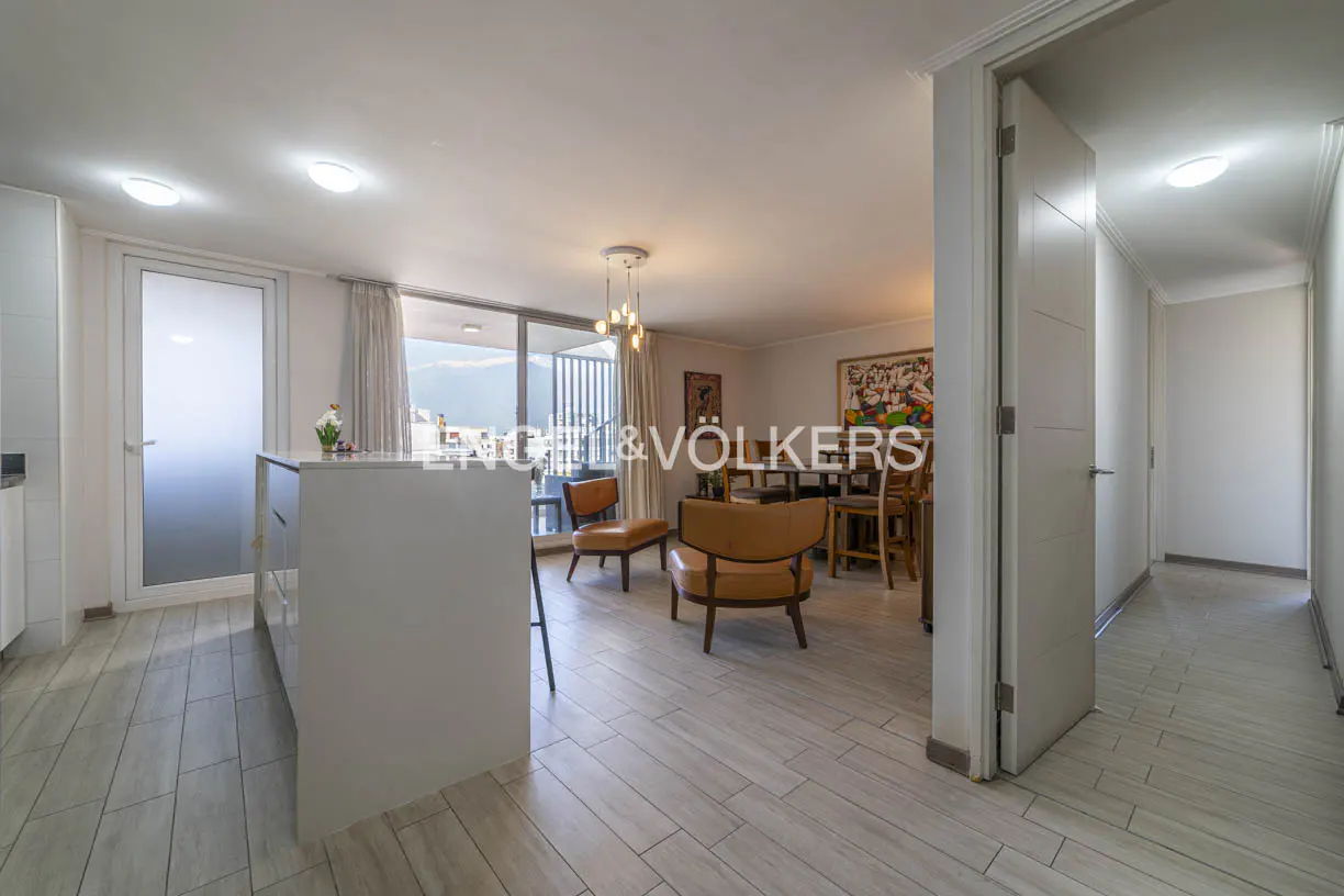 Bright, open-concept apartment with wood-look tile flooring, tan leather chairs, and a white kitchen island. A hallway leads to the right.
