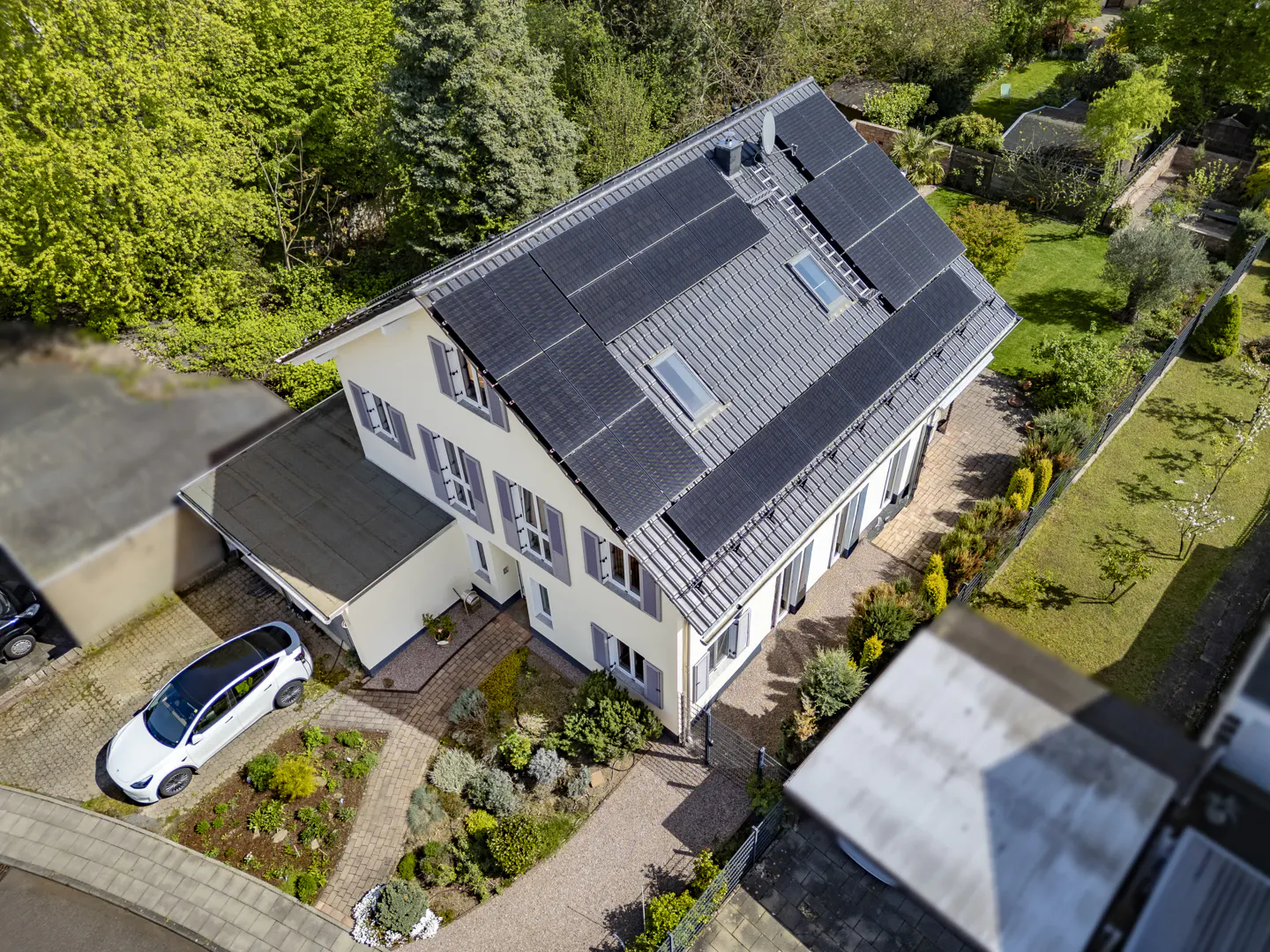 Aerial view of a two-story cream-colored house with gray shutters and a roof with solar panels, surrounded by green trees and a white car parked in the driveway.