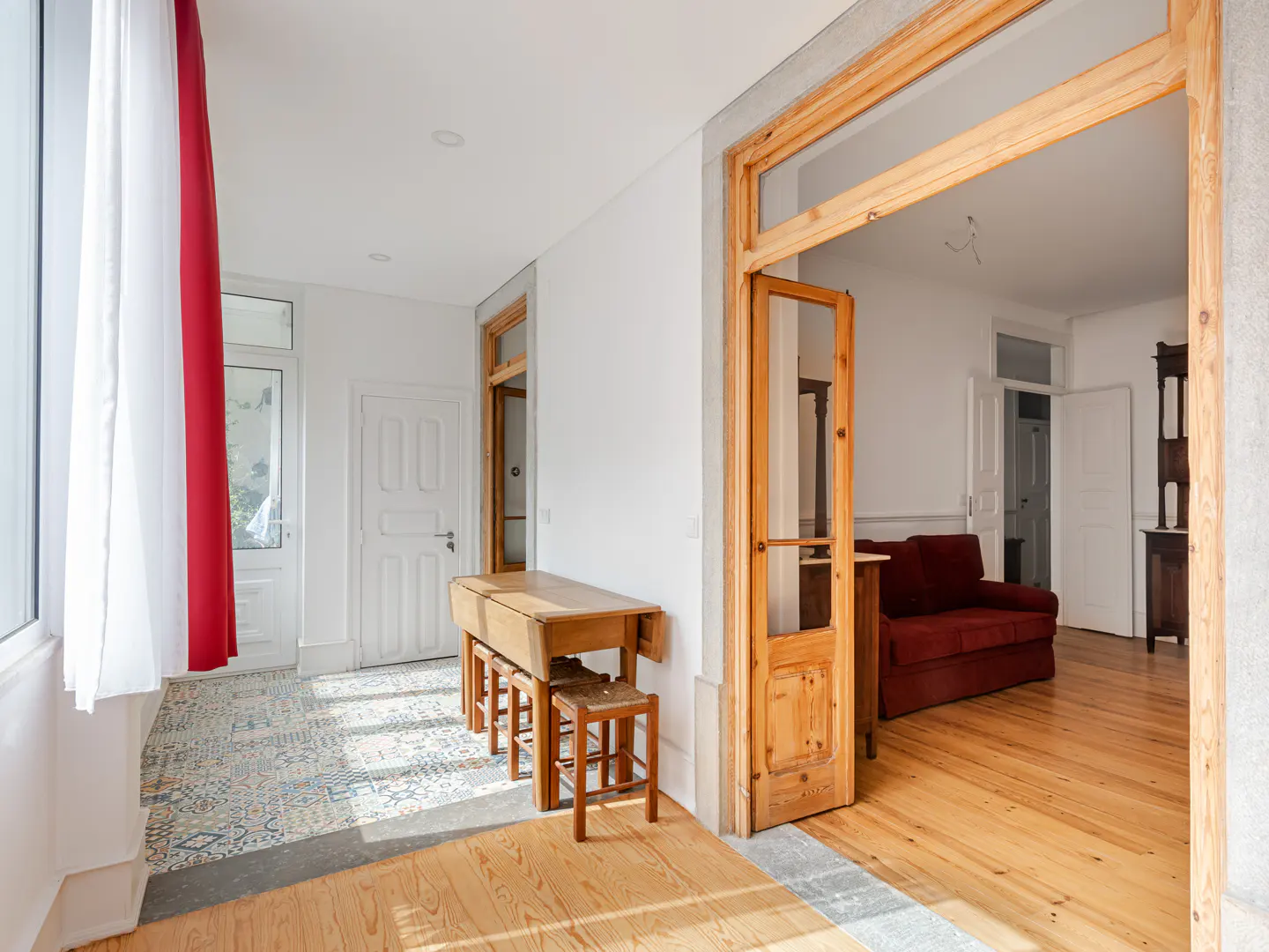 Bright hallway with patterned tile floor, wooden table and stools, leading to a living room with a red sofa and wood floors.