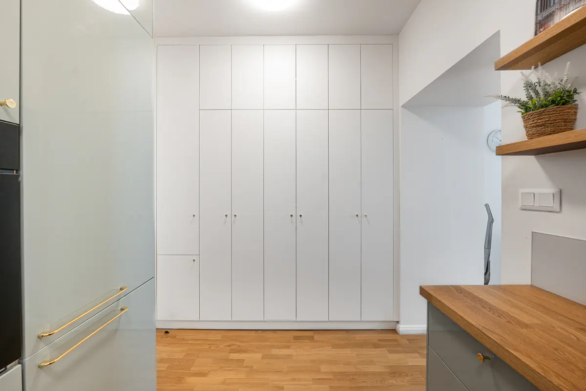Modern kitchen with floor-to-ceiling white cabinets, light wood flooring, and wooden shelves with a potted plant.
