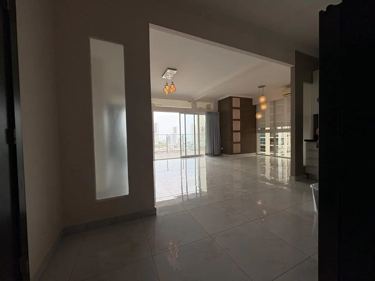 A bright, empty apartment with white tile floors, a balcony with a city view, and modern light fixtures.