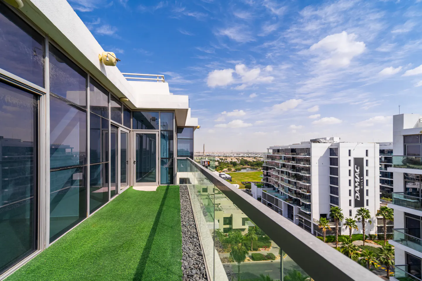 Balcony view with artificial grass, glass railing, and city skyline. "DAMAC Hills" building visible in the background. Blue sky with clouds.