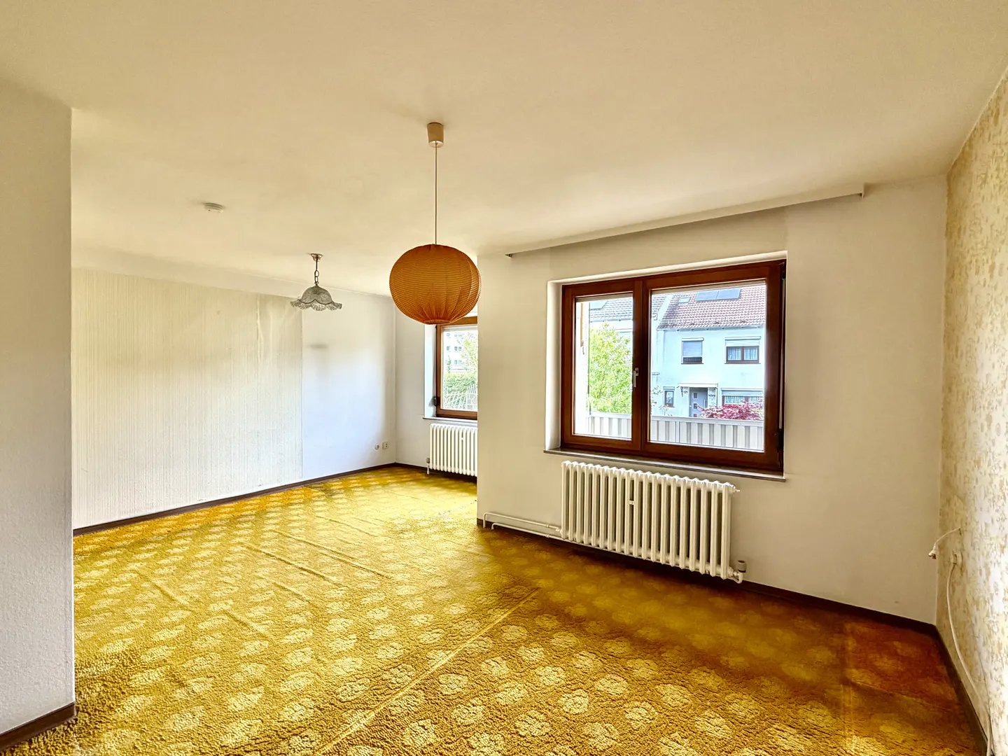 A vacant room with yellow patterned carpet, white walls, and two windows. A round orange pendant light hangs from the ceiling.