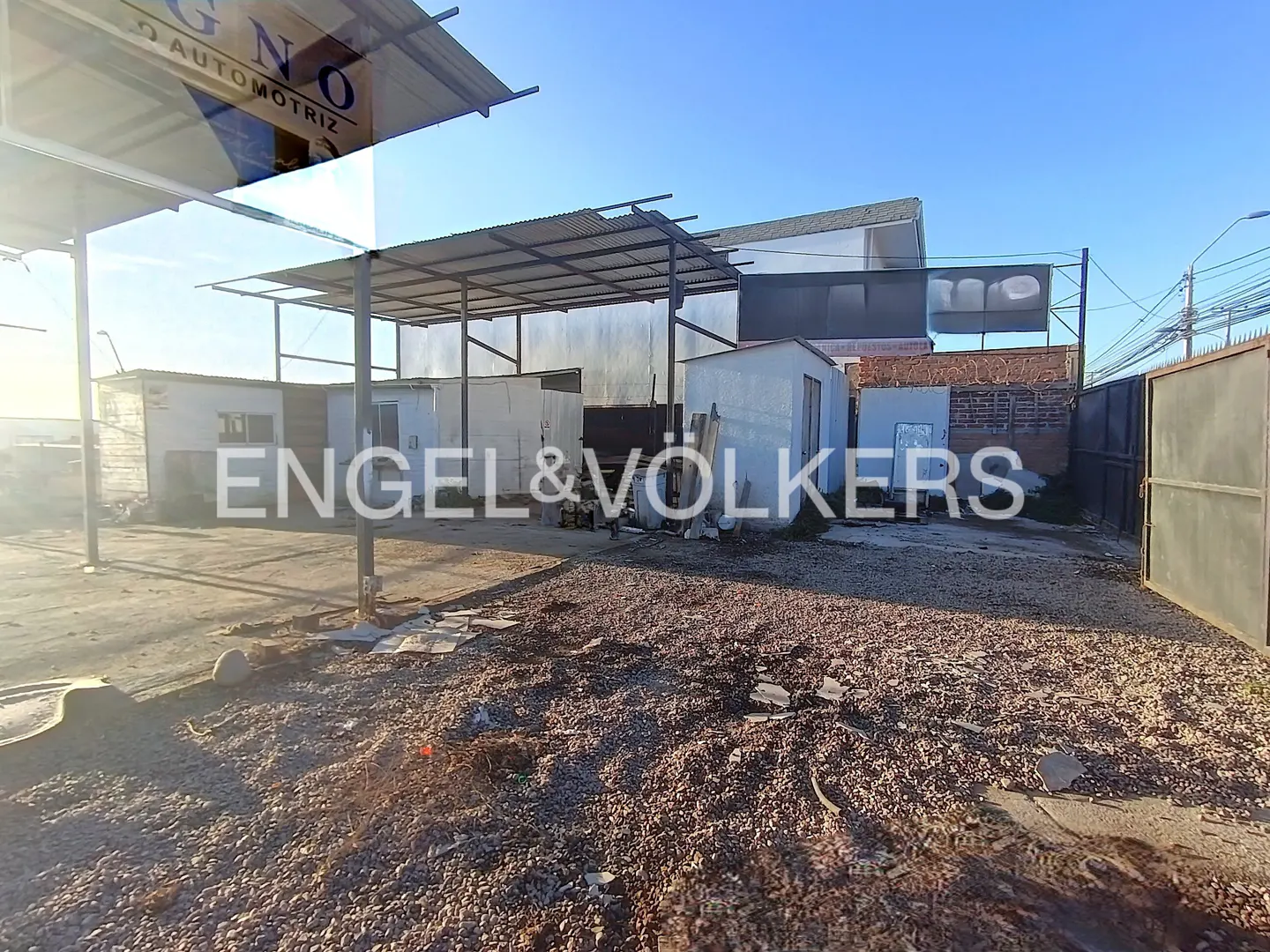 A gravel lot with small white buildings and metal awnings under a blue sky. The Engel & Völkers logo is superimposed on the image.