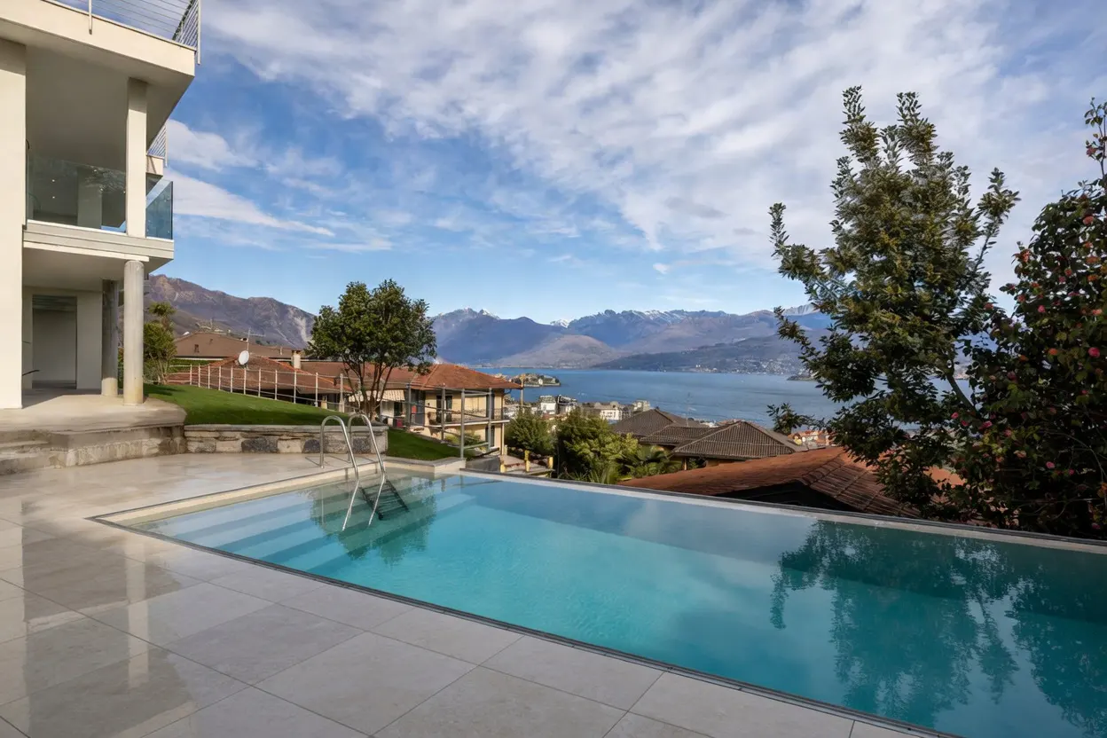Infinity pool with blue water reflects the sky, overlooking a lake and mountains. Trees and houses are in the background.
