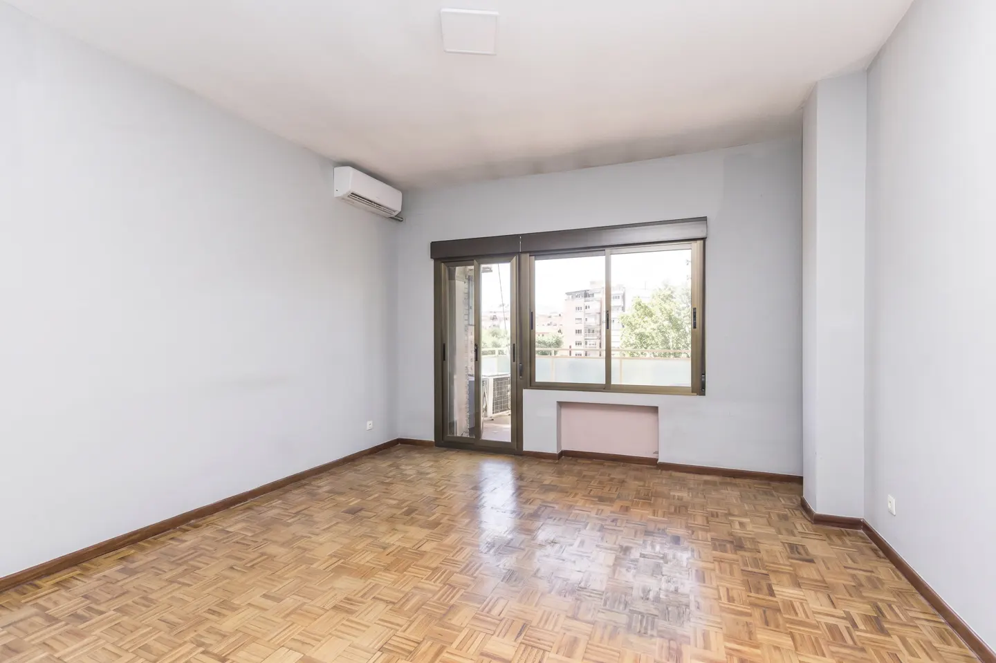 Empty room with light grey walls, parquet flooring, air conditioning unit, and a sliding glass door leading to a balcony.