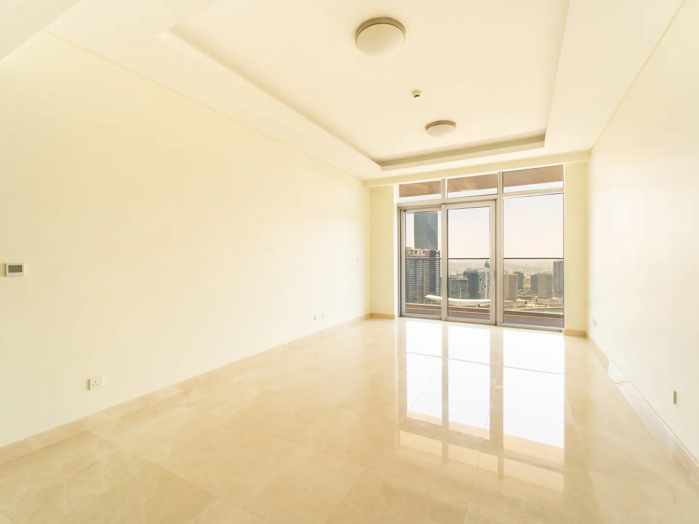 Empty, bright room with cream walls, polished tile floors, and floor-to-ceiling windows overlooking a city skyline.
