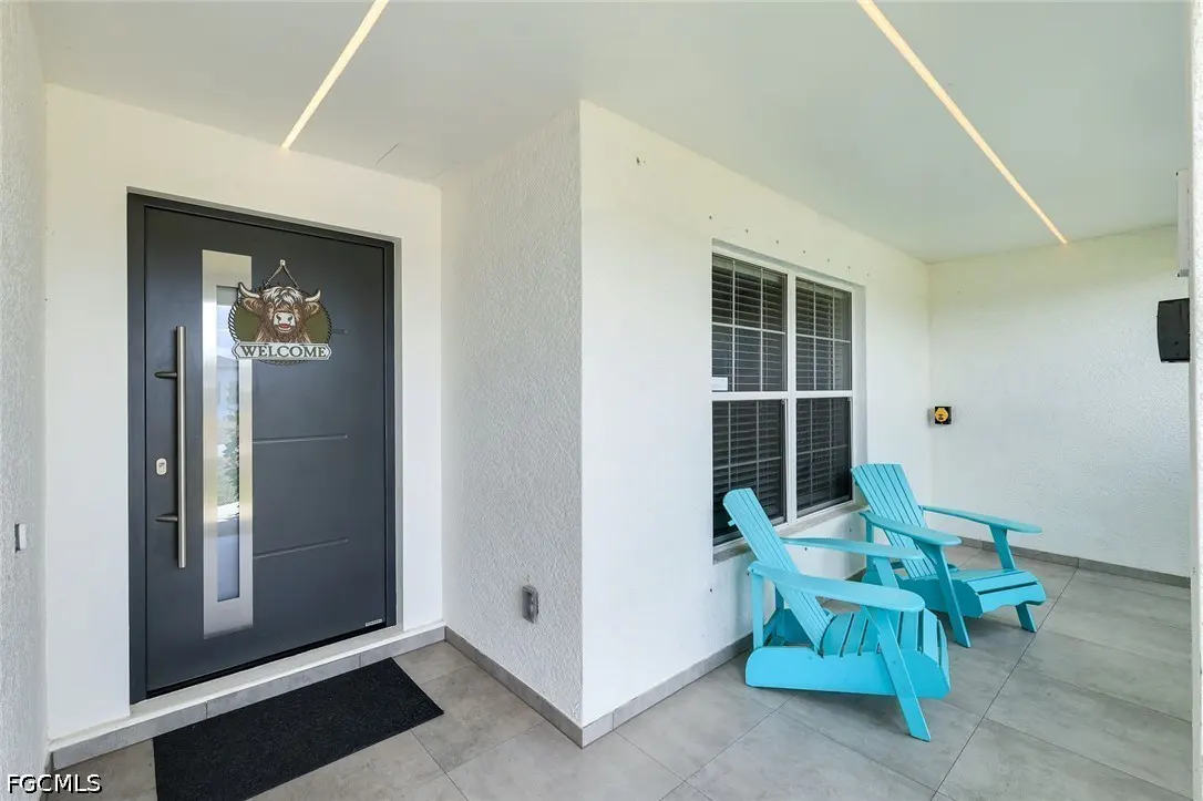 Modern home entrance with a gray door, "Welcome" sign, and two blue Adirondack chairs on a tiled porch.