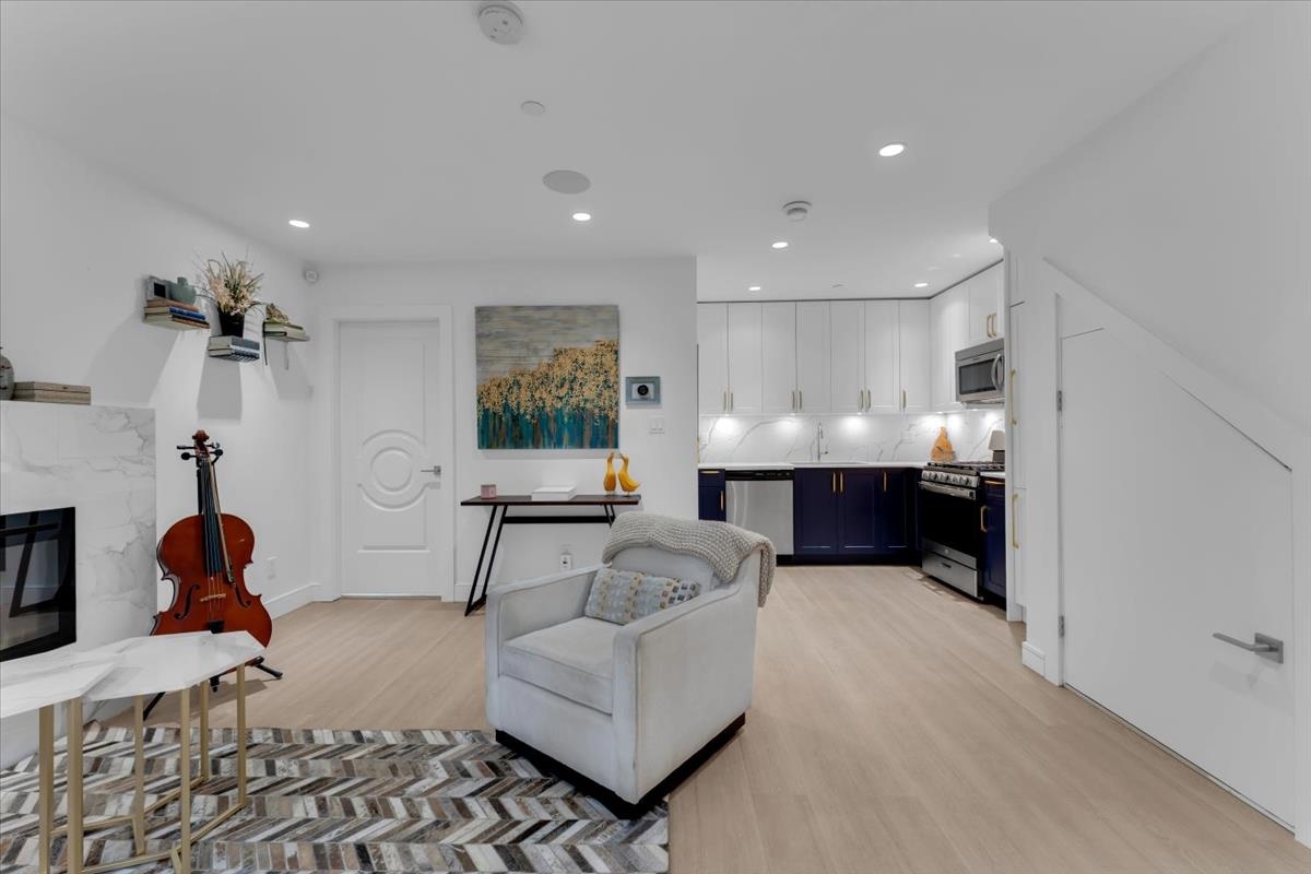 Bright living space with a white armchair, herringbone rug, and cello near a fireplace. A modern kitchen with white cabinets and blue lowers is visible.