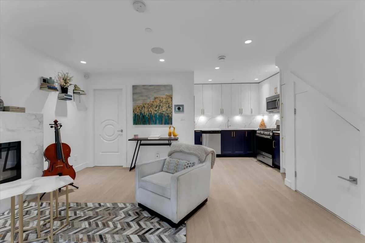 Bright living space with a white armchair, herringbone rug, and cello near a fireplace. A modern kitchen with white cabinets and blue lowers is visible.