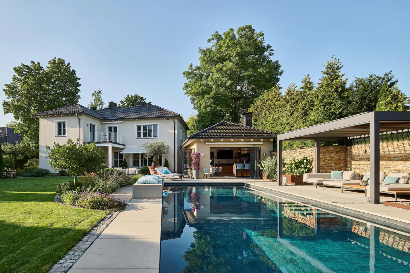 A backyard with a pool, a beige house, a pool house, and a pergola with outdoor furniture.