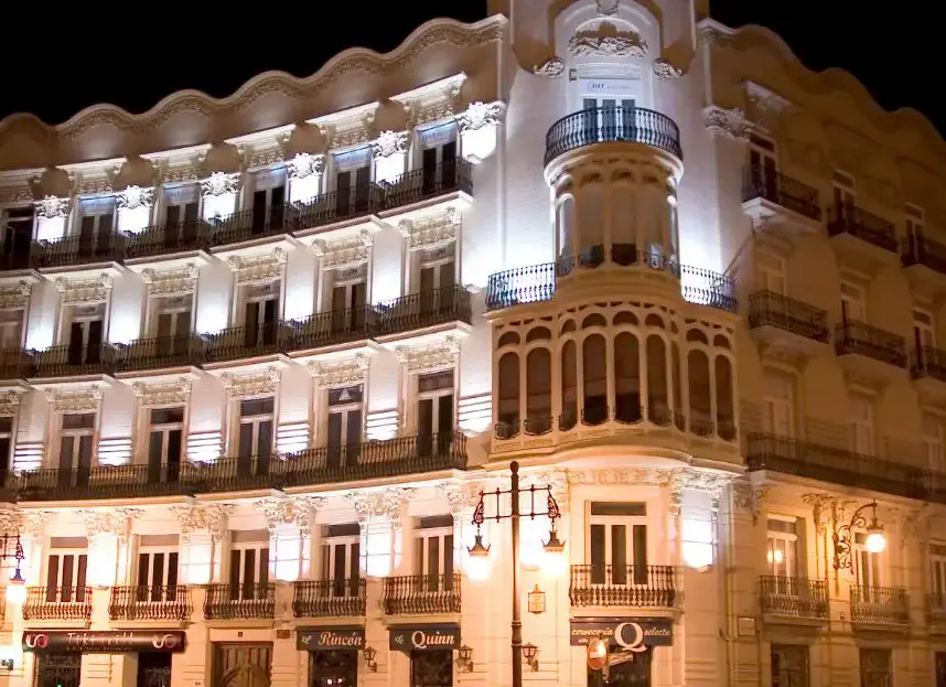 Night view of a multi-story white building with ornate balconies and windows, illuminated by streetlights. Businesses are visible on the ground floor.