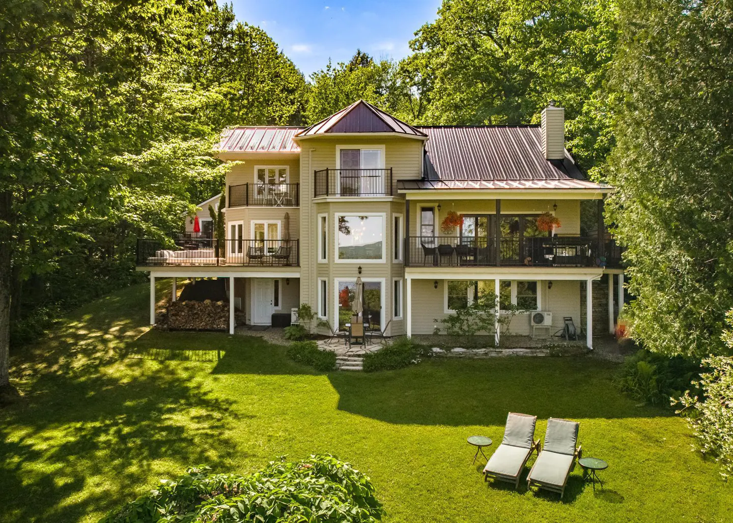 Two-story beige house with black metal roof and balconies, surrounded by green trees and lawn with two lounge chairs.