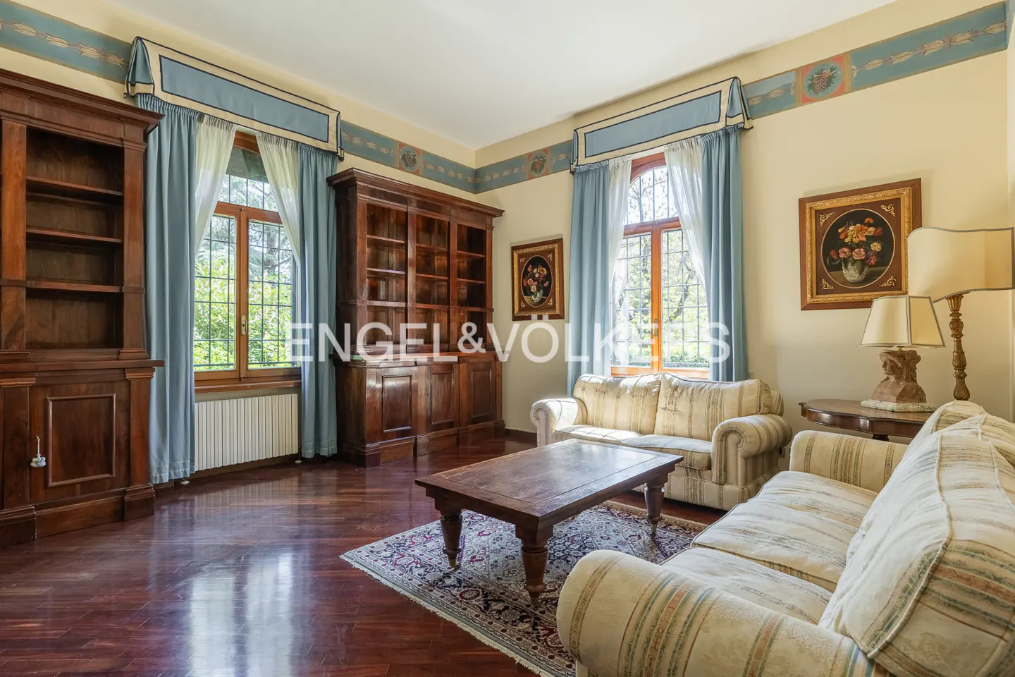 A living room with hardwood floors, two sofas, a wooden coffee table, and two wooden bookshelves. Two windows with blue curtains are visible.