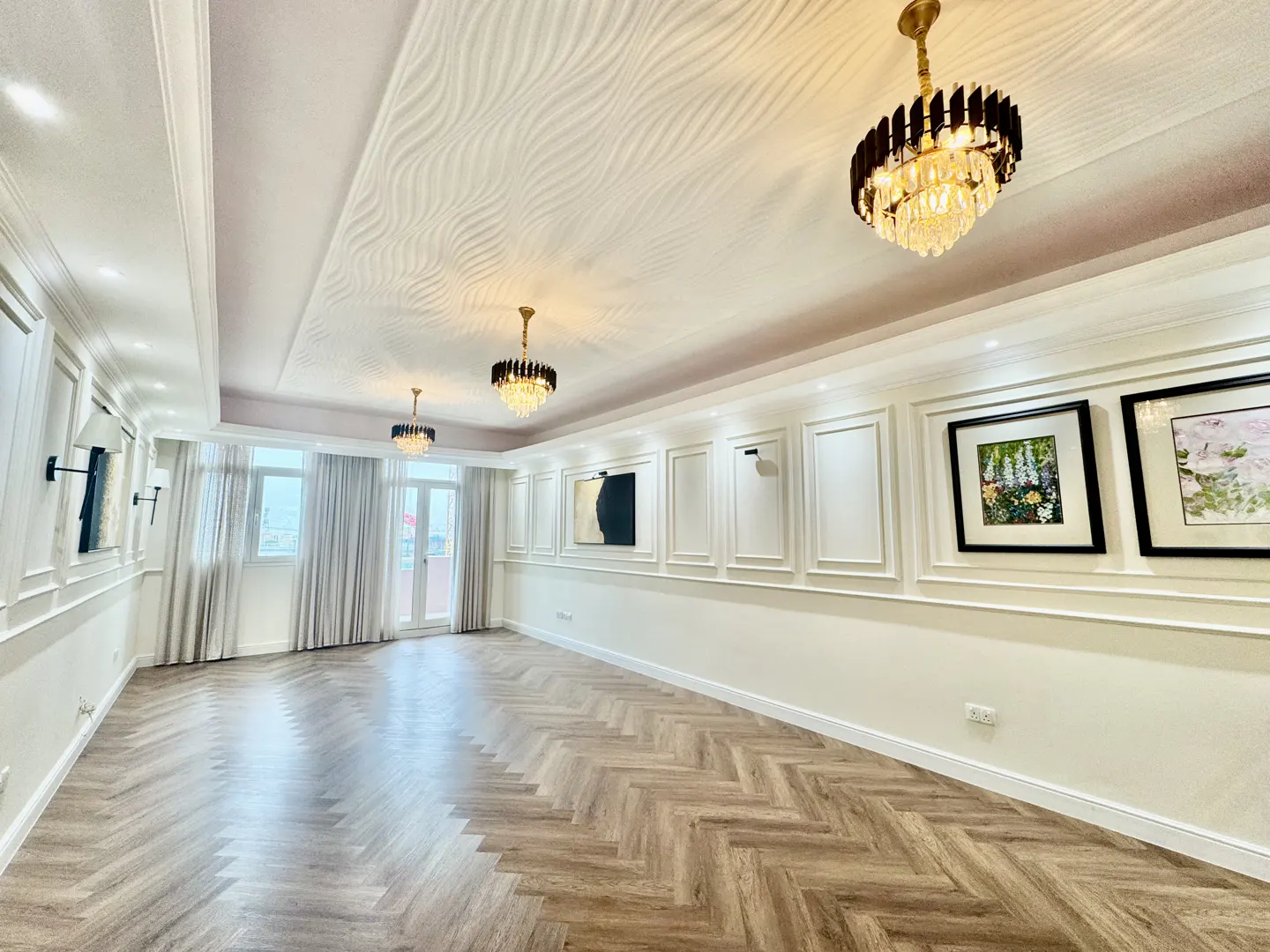 Bright, empty room with herringbone wood floors, white walls with molding, and three crystal chandeliers. Two framed pictures hang on the wall.