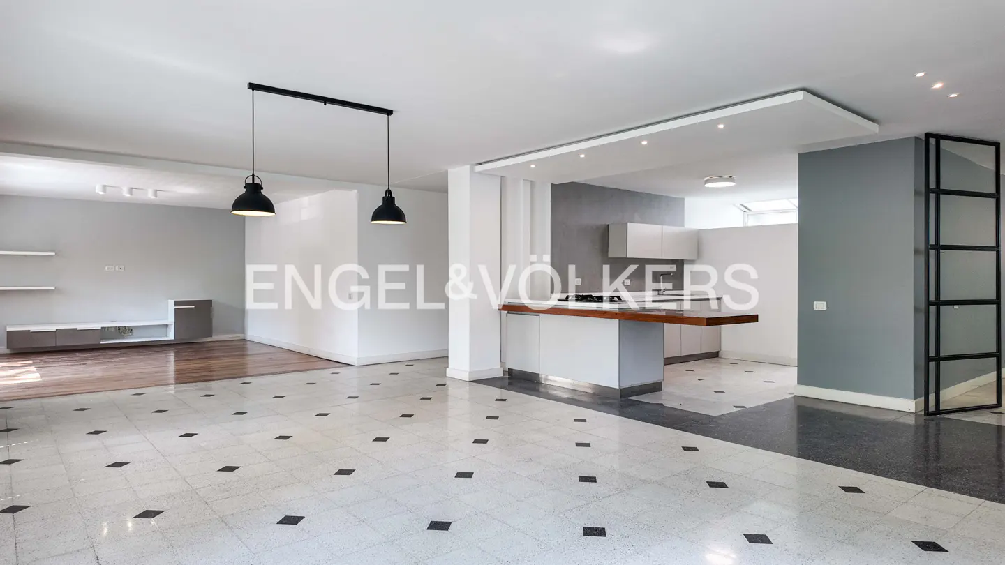 Open-concept living space with white walls, black pendant lights, and a kitchen island with a wood countertop. The floor is tiled with black squares.