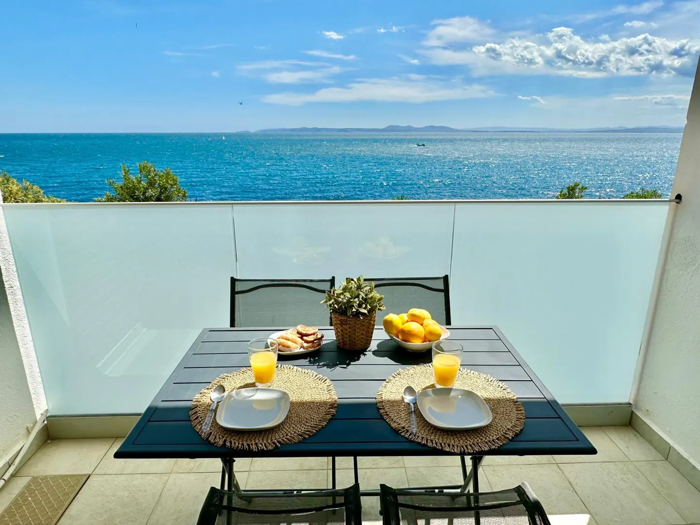 Balcony view with breakfast table overlooking the ocean. Table set with plates, juice, fruit, and pastries. Blue sky and water.