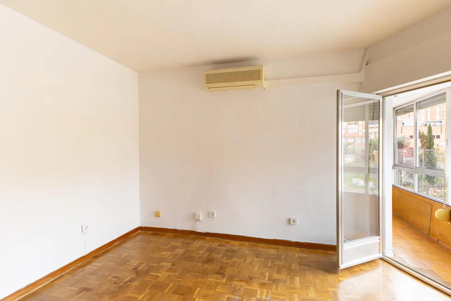 Bright, empty room with parquet floor, white walls, and an open glass door to a balcony with a city view.