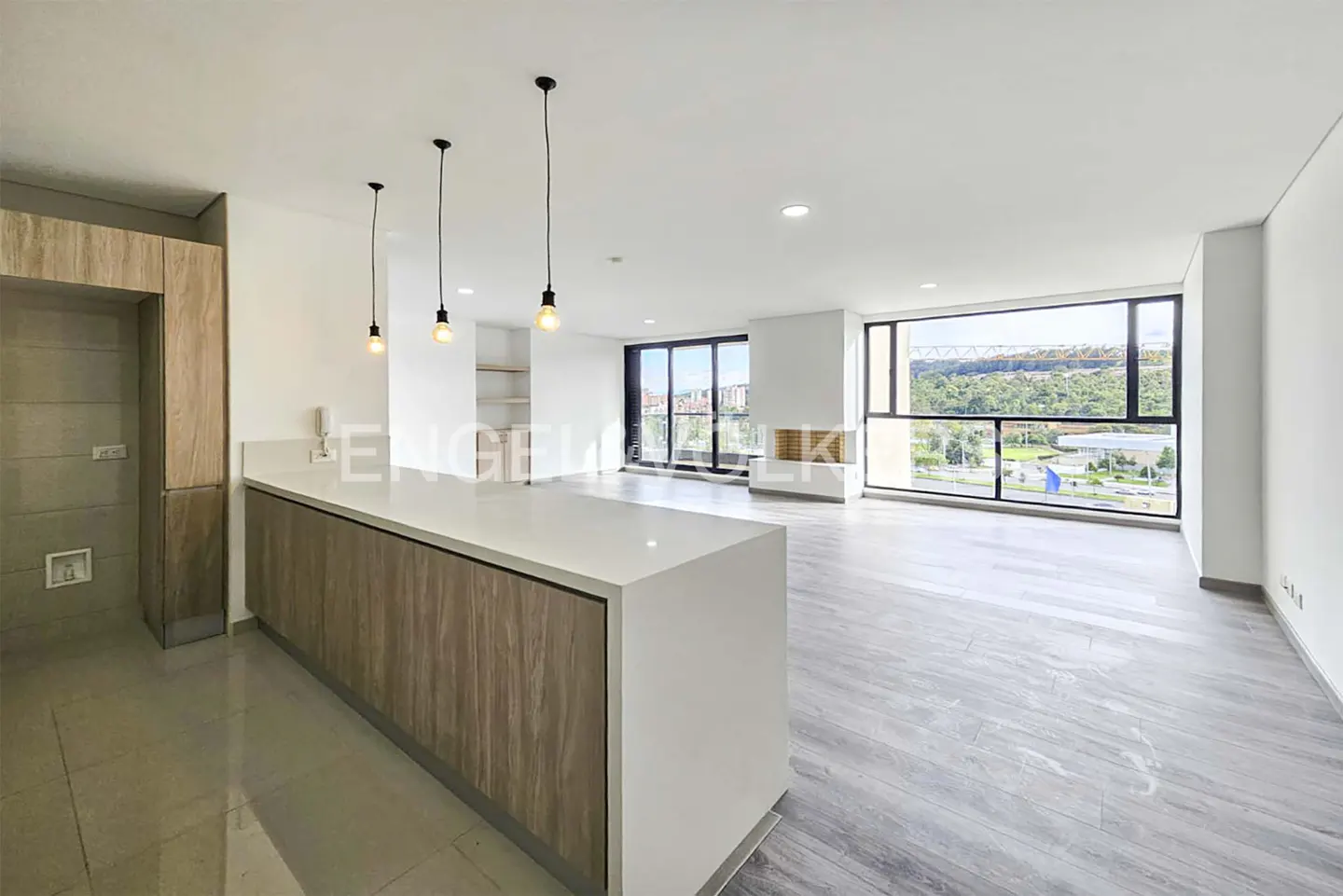 Modern open-plan kitchen with wood cabinets, white island, pendant lights, and large windows overlooking a green landscape.
