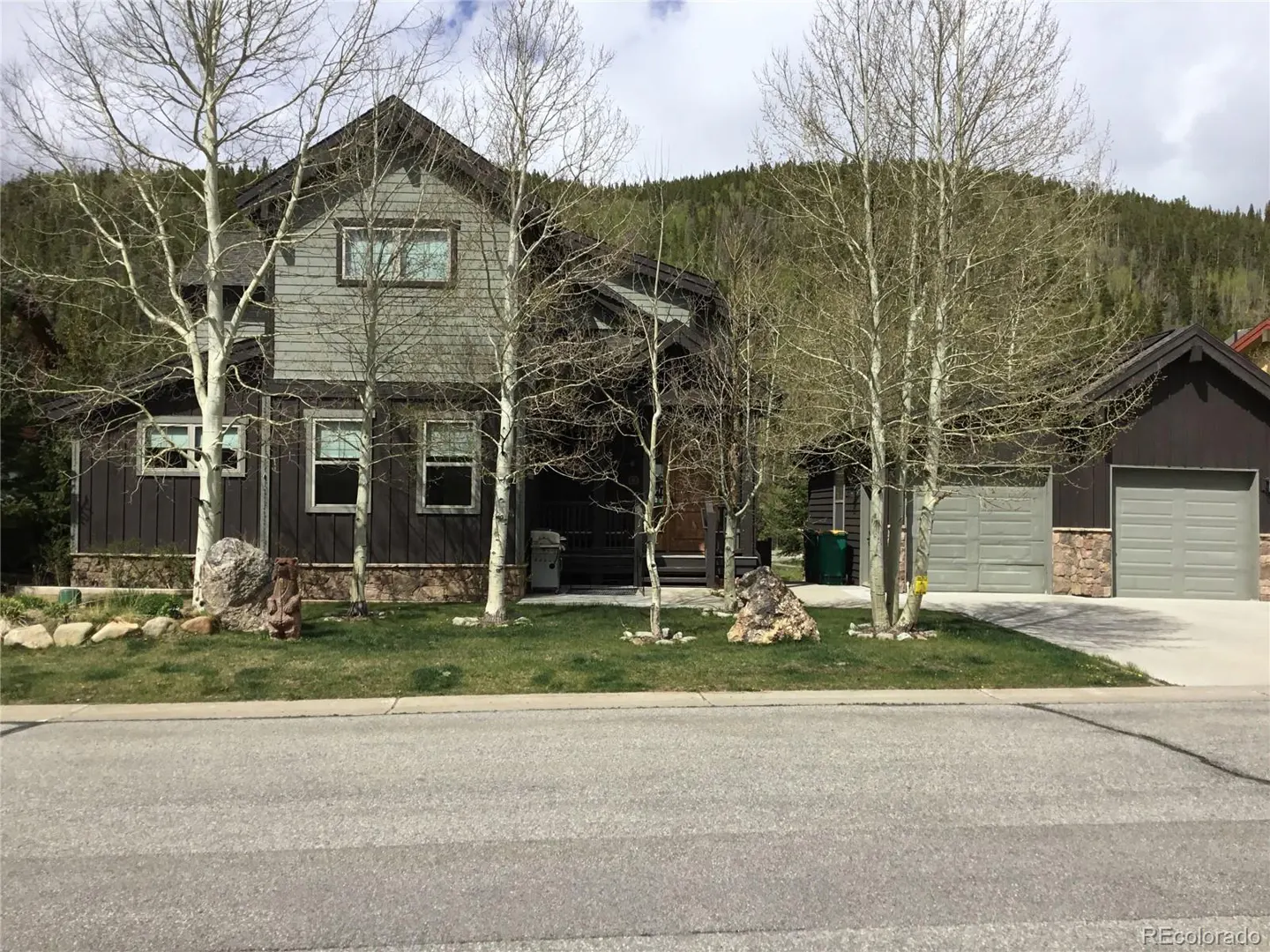 Two-story house with gray siding and a brown base, surrounded by bare trees and a green lawn. A gray garage is on the right.