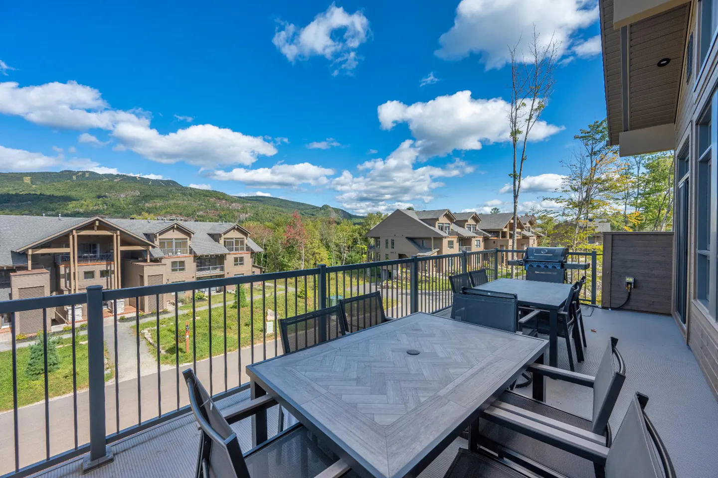 Balcony view with tables, chairs, and grill. Mountains and blue sky with clouds in the background. Railing in the foreground.