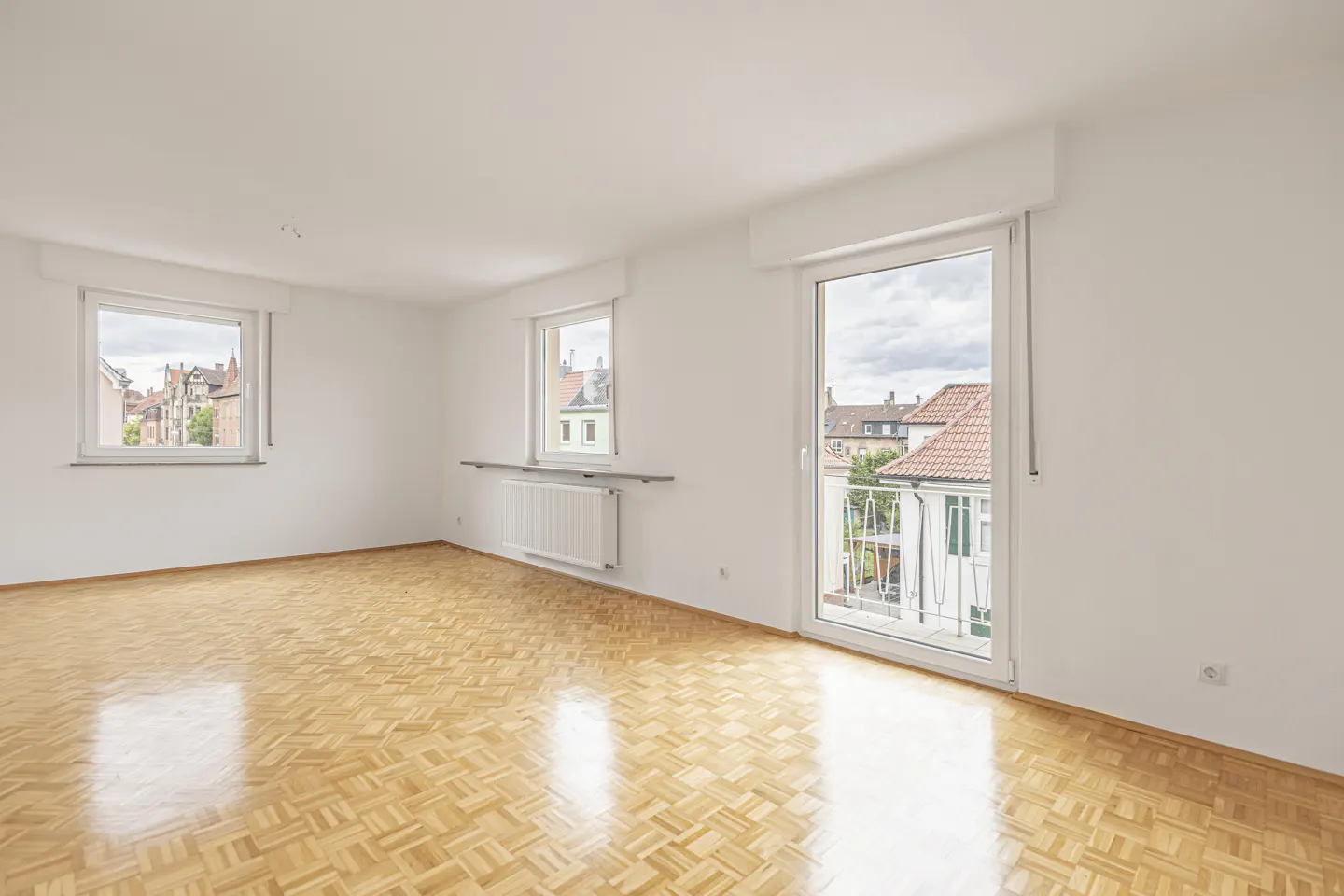 Bright, empty room with parquet floor, white walls, two windows, and a balcony door showing a view of neighboring houses.